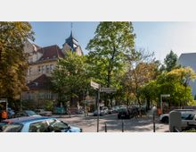 Straßenszene mit dichter Begrünung und einem historischen Gebäude mit rotem Ziegeldach und Turm im Hintergrund. Straßenschilder mit den Namen "Schwalbacher Straße" und "Rheingaustraße" stehen in der Mitte; parkende Autos säumen die Straße.