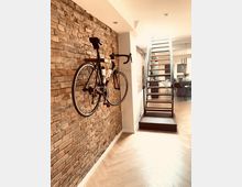 Ein modernes, offenes Wohnraumdesign mit einer freigelegten Ziegelwand, an der ein Fahrrad vertikal aufgehängt ist. Rechts befindet sich eine schmale, metallische Treppe, die in ein höheres Stockwerk führt, und dahinter ein Essbereich mit hängenden Lampen und einem gedeckten Tisch.