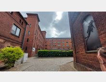 Backstein-Gebäudekomplex mit mehreren Stockwerken, umgeben von einem gepflasterten Innenhof und grünen Büschen; an der rechten Gebäudewand befindet sich ein großes Fotoposter mit einem Motiv eines Tauchers.