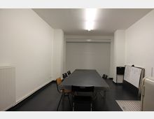 Schlichter Raum mit weißen Wänden, schwarzem Boden und einer langen rechteckigen Tischgruppe, umgeben von einfachen Stühlen. Im Hintergrund befindet sich ein geschlossenes Rolltor, seitlich stehen eine Tafel und ein Heizkörper.