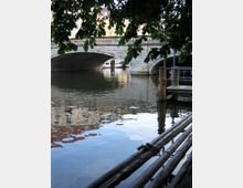 Steinerne Brücke über einen Fluss mit Bäumen im Vordergrund, einem kleinen Holzsteg und Spiegelungen von Gebäuden im Wasser. Im Hintergrund ist ein Boot unter der Brücke sichtbar.