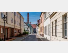 Schmale gepflasterte Straße in einer Altstadt mit zweistöckigen Wohnhäusern auf beiden Seiten, darunter ein Fachwerkhaus. Im Hintergrund ragt ein roter Schornstein empor, und Laternen im historischen Stil säumen den Gehweg.