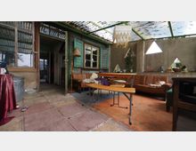 Ein rustikaler Innenraum mit sichtbaren Holzwänden und Wellblechdach, möbliert mit einem Holztisch, Bänken und einer Ledercouch. Fenster unterschiedlicher Form lassen Tageslicht herein, während der Boden aus unregelmäßigen Steinplatten besteht.