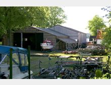 Ein Werftgelände mit einem großen Schuppen aus Holz und Wellblech, in dem ein Boot steht, das teilweise aus dem offenen Eingang herausragt. Im Vordergrund liegen Anhänger und verschiedene Metallgestelle, während im Hintergrund Bäume und ein Blick auf ein Gewässer zu sehen sind.