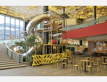 Innenbereich eines Freizeit- oder Einkaufszentrums mit einer großen Indoor-Rutsche und einem Klettergerüst aus Metall und Holz, umgeben von gelben Sicherheitsabsperrungen. Im Vordergrund stehen Tische und Stühle, während links eine Treppe zu einer oberen Ebene mit großen Fenstern führt.