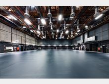 Großer, leerer Industriebereich mit grauem Boden, schwarzen Wänden und einer hohen Decke mit sichtbaren Rohren, Lüftungsschächten und Metallträgern. Die Beleuchtung erfolgt durch helle, rechteckige Lampen, die an der Decke befestigt sind.