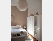 Minimalist bedroom with a taupe accent wall, white bed linens, and a large round paper pendant light hanging from a high ceiling with molding. Along one wall are tall white wardrobes with paneled doors and open shelving, set on warm wooden floors beside a small bedside table and lamp.