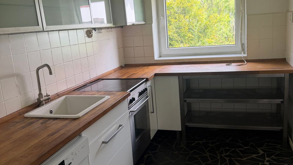 Kleine Küche mit L-förmiger Holzarbeitsplatte, weißer Spüle, Herd und weißen Unterschränken sowie Hängeschränken mit Glasfronten. Ein großes Fenster mit Rollladen bringt Tageslicht hinein; weiße Fliesenrückwand und dunkler, unregelmäßig gemusterter Steinboden.