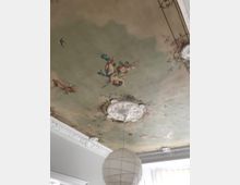 Detailreiche Zimmerdecke mit einem Deckenfresko, das Engel und florale Ornamente zeigt, umrahmt von weißen Stuckverzierungen. Im Vordergrund hängt eine schlichte, weiße Papierlampenschirm von der Decke.