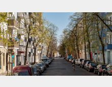 Straßenansicht mit mehrstöckigen Wohnhäusern auf beiden Seiten, gesäumt von Bäumen mit jungen, grünenden Blättern. Parkende Autos reihen sich entlang der Fahrbahn, und eine Person fährt mit dem Fahrrad auf der Straße.