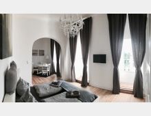 Helles Schlafzimmer mit einem Doppelbett und grauer Bettwäsche, Holzboden, einem Kronleuchter und bodenlangen dunklen Vorhängen vor großen Fenstern. Im Hintergrund ist ein Rundbogen, durch den man einen Essbereich mit Tisch und Stühlen sehen kann.