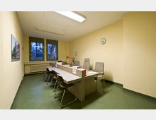 Büro mit einem langen Empfangstresen, mehreren Stühlen auf beiden Seiten, hellgelben Wänden, zwei Fenstern, einer Wanduhr und einem Bild an der Wand. Der Boden ist mit grünem Linoleum ausgelegt und durch Deckenleuchten beleuchtet.