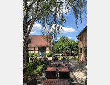 Ein Außenbereich mit Tischen und Stühlen aus dunklem Holz und Korbgeflecht, umgeben von Bäumen und einem gepflasterten Boden. Im Hintergrund ein Fachwerkgebäude und ein Backsteinhaus, umgeben von grüner Vegetation und Menschen, die den sonnigen Tag genießen.