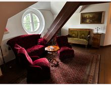 Wohnzimmer im Vintage-Stil mit roten Polstermöbeln, einem kleinen Holztisch und einem großen, runden Fenster. Holztragbalken und ein Teppich mit orientalischem Muster prägen den Raum, daneben befinden sich ein antikes Sofa, ein Schrank und ein Gemälde an der Wand.