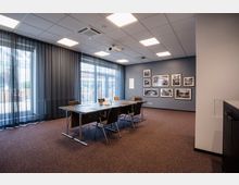 Ein moderner Konferenzraum mit einem rechteckigen Tisch und mehreren Stühlen auf Teppichboden. Die Wände sind mit blauen Akzenten gestrichen und mit gerahmten Schwarz-Weiß-Fotografien dekoriert; große Fenster mit halbtransparenten Vorhängen lassen Tageslicht herein.