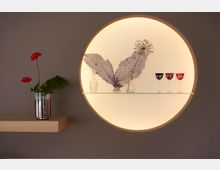 Rundes Wandregal mit Hintergrundbeleuchtung, das auf Glas dekorative Objekte wie getrocknete Pflanzen, kleine Flaschen und farbige Weingläser zeigt; daneben ein schlichtes Holzregal mit einer silbernen Vase und einer roten Blume.