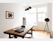 Heller Raum mit einem großen Holztisch und Hockern auf einem Holzboden, große Fensterfront mit Tür, eine Stehlampe rechts und ein Spiegel im Hintergrund; an der Wand hängt ein Bild mit goldener Blüte.