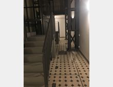 Treppenhaus mit neutralem Licht, einer gemusterten Fliesenbodenfläche in Schwarz-Weiß und Metallgeländern. Im Hintergrund sind metallene Tragstrukturen und Wände aus Backstein sowie ein Aufzugsschacht erkennbar.