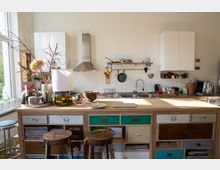 Helle, moderne Wohnküche mit einer Holzinsel im Vordergrund, umgeben von Barhockern. Im Hintergrund befinden sich ein Waschbecken mit Armaturen, weiße Hängeschränke, ein Wandregal mit Kochutensilien und eine Dunstabzugshaube; dekorative Gegenstände und Pflanzen sind auf der Insel arrangiert.