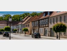 Straßenzug in einer Kleinstadt mit Kopfsteinpflaster, parkenden Autos und kugelförmig beschnittenen Straßenbäumen. Entlang der Straße stehen eng gereihte, zweigeschossige Wohnhäuser mit Putzfassaden und teils Fachwerkdetails unter roten Satteldächern.