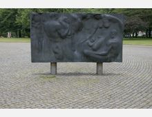 Bronzeflügel mit Reliefdarstellung von menschlichen Figuren auf einem gepflasterten Platz, umgeben von Rasenflächen und Bäumen im Hintergrund. Die Skulptur steht auf kurzen Zylindersäulen.