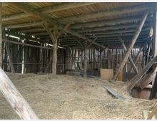 Holzscheune mit offenem, sichtbarem Gebälk und Holzdach; der Boden ist mit Heu bedeckt, und die Wände bestehen aus vertikalen Holzplanken mit Lücken, durch die Licht einfällt.