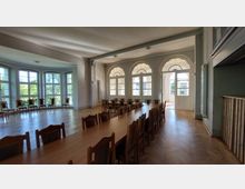 Großer, heller Versammlungsraum mit Parkettboden, langen Holztischen und Stuhlreihen. Im Hintergrund befinden sich große Fenster und drei Rundbogen-Türen, die auf eine Terrasse oder einen Außenbereich führen.