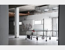 Industriehalle mit Betonwänden und sichtbaren Rohrleitungen, ausgestattet mit einer Tischtennisplatte in der Mitte des Raumes. An der Wand hängt ein abstraktes Gemälde, und durch kleine Fenster fällt Tageslicht.