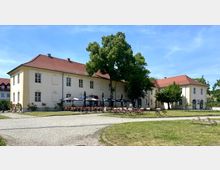 Historisches zweistöckiges Gebäude mit hellgelber Fassade und rot gedecktem Walmdach, umgeben von einem weitläufigen Garten mit Rasenflächen. Vor dem Gebäude befinden sich Sitzmöglichkeiten eines Außencafés, teils mit Sonnenschirmen, sowie vereinzelte große Bäume.