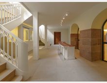 Innenansicht eines hellen, modern renovierten Gebäudes mit weißen Wänden und geschwungenen Treppen mit dekorativem Geländer. Der Raum verfügt über einen roten Sandsteinsockel an den Wänden, einen zentral platzierten Tisch und eine Holzrahmentür im Hintergrund.