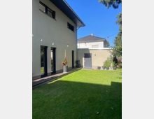 Moderne Hausfassade mit beigem Putz und großen schwarzen Fenstern, die zu einer Holzterrasse führen. Vor dem Haus befindet sich ein gepflegter Rasen, umgeben von vereinzelten Pflanzen in Töpfen und Bäumen im Hintergrund.