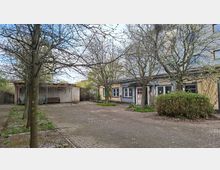 Innenhof mit gepflasterter Fläche und mehreren kahlen Bäumen, umgeben von niedrigen Gebäudeteilen mit Fenstern und einer Tür mit farbigem Rahmen. Links steht ein offener Unterstand mit Sitzbank, rechts sind Sträucher und kleine Grünflächen am Rand zu sehen.