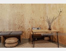 Minimalistisch eingerichteter Raum mit Wänden aus hellem Sperrholz. Zu sehen sind ein kleiner Holztisch mit einer Glasvase und Zweigen, eine Sitzbank mit grauen Polstern sowie runde, geflochtene Sitzkissen auf einem Teppich aus Naturfasern.