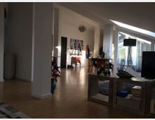 Großer, moderner Wohnraum mit Holzboden, weißen Wänden und großen Fenstern, die viel Tageslicht hereinlassen. Der Raum ist mit minimalistischen Möbeln, dekorativen Elementen und einem Teppich gestaltet, im Hintergrund ist eine Tür und eine abstrakte Wanddekoration erkennbar.