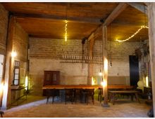 Großer, rustikaler Raum mit unverputzten Backsteinmauern, Holzdecke und sichtbaren Holzbalken. Ein langer Holztisch mit Stühlen steht zentral, umgeben von warmem Licht und einer dekorativen Lichterkette; im Hintergrund sind ein alter Schrank und ein hängendes Fahrrad sichtbar.