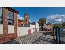 Gepflasterte Straße in einer kleinen Ortschaft mit historischen Gebäuden, einer modernen Fassade links und einem Kirchturm mit Spitzdach im Hintergrund. Der Bereich ist von Bäumen und Wohnhäusern umgeben, und das Wetter ist sonnig mit vereinzelten Wolken.