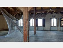 Ein historischer Innenraum mit Holzbalkendecke, freiliegenden Backsteinwänden und großen, bogenförmigen Fenstern. Im Raum ist eine filigrane Kunstinstallation aus gespannten weißen Fäden zwischen den Holzbalken zu sehen.