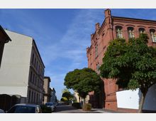 Straßenszene mit einem historischen, roten Backsteingebäude auf der rechten Seite und einem hell verputzten Wohngebäude links. Entlang der Straße stehen geparkte Autos und mehrere Bäume, die einen schmalen Gehweg beschatten.