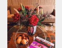 Ein Glastisch mit einem silbernen Metallbehälter, der rote und lilafarbene Blumen enthält, steht im Vordergrund. Daneben liegen eine Schale mit Früchten und ein Albumcover mit der Aufschrift "Frank Zappa"; im Hintergrund befindet sich ein beiges Sofa und eine Wand mit vertikalen Holzpaneelen.