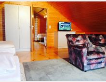 Wohnzimmer mit Holzverkleidung in einem Dachgeschoss, ausgestattet mit einem Polstersessel, einem TV auf einer Kommode, einem Kleiderschrank und sichtbaren Betten. Eine offene Tür führt in einen angrenzenden Raum mit weiteren Möbeln.