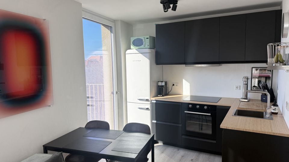Moderne, kompakte Wohnküche mit schwarzen Hängeschränken, Holzarbeitsplatte, Herd mit Backofen und Spülbecken sowie freistehendem Retro-Kühlschrank. Daneben ein Esstisch mit Stühlen vor einer großen Balkontür, die viel Tageslicht hineinlässt.