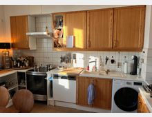 Küche mit Holzfronten, weißen Fliesen und integrierten Geräten. Sichtbar sind ein Herd, eine Spülmaschine, eine Waschmaschine, eine Spüle und Arbeitsflächen mit Küchenutensilien, sowie Regale und Hängemessern an der Wand.