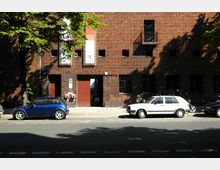 Backsteinfassade eines Gebäudes mit kleinen quadratischen Fenstern und einem roten Doppeltor als Eingang. Vor dem Gebäude parken mehrere Fahrzeuge, darunter ein blauer und ein weißer Kleinwagen, sowie zwei Motorräder; im Vordergrund eine Straße mit einem Fahrradstreifen.