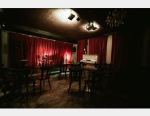 Small performance room with a low stage backed by deep red curtains, two chairs under stage lights, and musical instruments nearby. The space has wooden plank flooring, café-style tables and chairs facing the stage, a white upright piano, and ceiling-mounted lights with a chandelier.