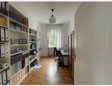 Ein kleiner, heller Arbeitsraum mit Holzdielenboden, einem Bücherregal voller Ordner und Dokumente auf der linken Seite und einem Schreibtisch mit Bürostuhl vor einem Fenster mit weißen Vorhängen. Rechts befindet sich ein Schrank mit Holzlamellentüren.