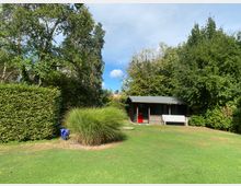 Holzhaus mit überdachter Veranda in einem gepflegten Garten, umgeben von hohen Bäumen und Sträuchern. Im Vordergrund eine Rasenfläche mit einer kleinen Hecke und Ziergrasbüscheln.
