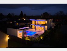 Nachtaufnahme eines modernen, zweigeschossigen Flachdachhauses mit großen Glasfronten und Balkon, angrenzend an eine beleuchtete Terrasse mit rechteckigem Swimmingpool. Das Anwesen ist eingezäunt und liegt in einem Wohngebiet mit umliegenden Häusern und Bäumen im Hintergrund.