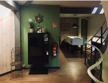 Gemütlicher Innenraum mit Holzboden, einer grünen Wand mit Deko wie einem Pflanzentopf und einem Schiffssteuerrad. Zu sehen sind ein Verkaufsautomat, ein Kickertisch im Hintergrund, eine Wendeltreppe rechts sowie Feuerlöscher an der Wand.