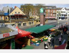 Straßenansicht mit einem kleinen Wochenmarkt, auf dem Obst und Gemüse unter grünen Markisen angeboten werden, daneben ein rotes Pizzastanddach. Im Hintergrund stehen mehrstöckige Häuser mit Geschäften und Restaurants, sowie ein S-Bahn-Schild auf einer gemauerten Säule.
