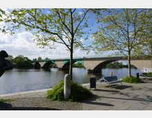 Eine Flusslandschaft mit einer steinernen Bogenbrücke, die den Fluss überspannt. Im Vordergrund befinden sich ein Gehweg mit Pflastersteinen, Sitzbänke, Bäume und ein Teil einer Skulptur.